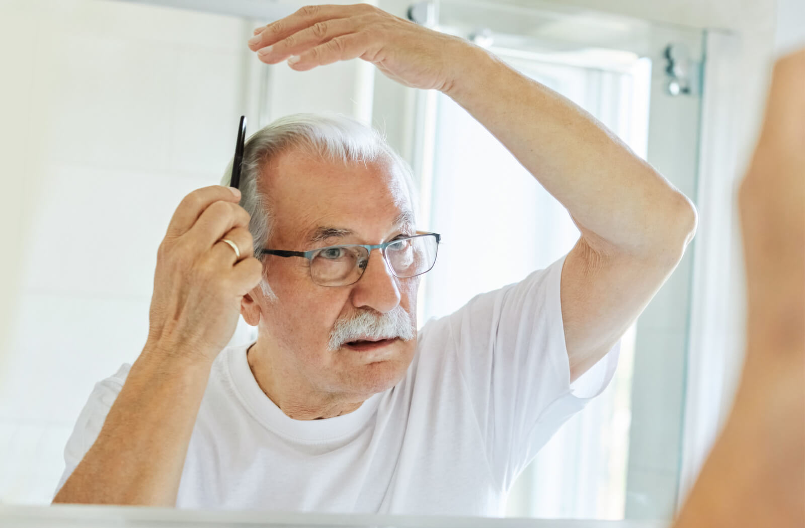 A senior man in a white shirt combing his hair in front of a mirror.