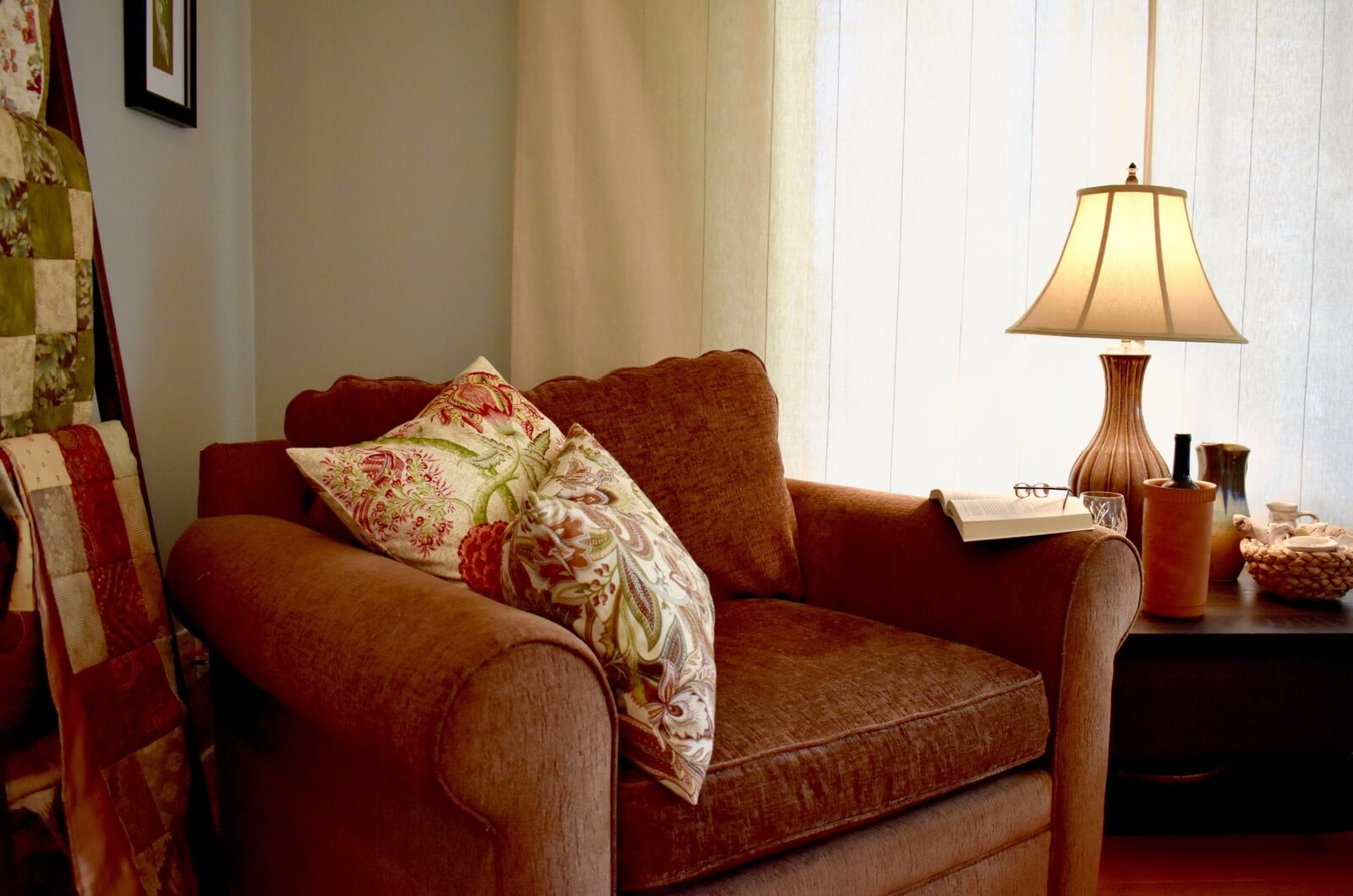 An image of a comfortable armchair with throw pillows next to a table with a lamp in a bright room.