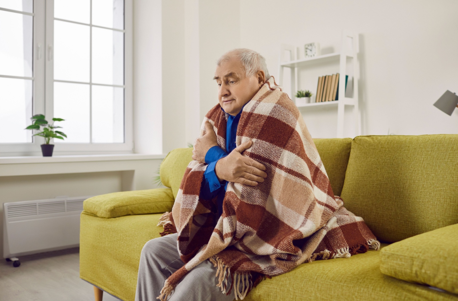 An older adult sitting on a couch trying to keep warm with a blanket wrapped around them.