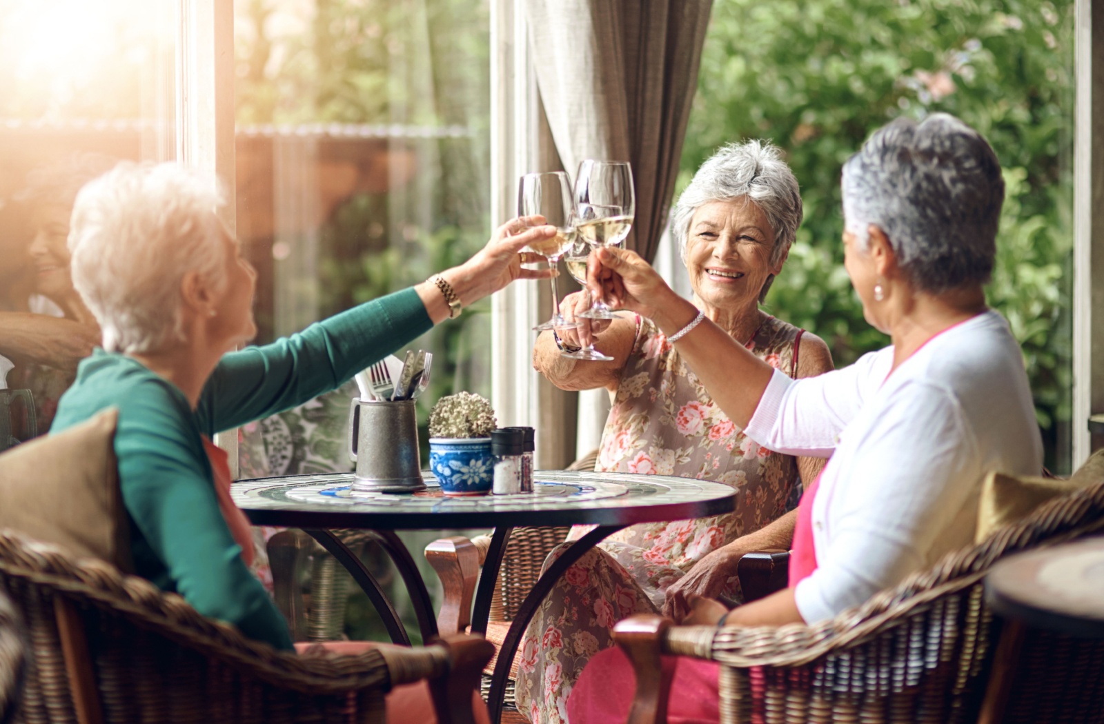 Three older friends sitting around a patio table, raising wine glasses and laughing.