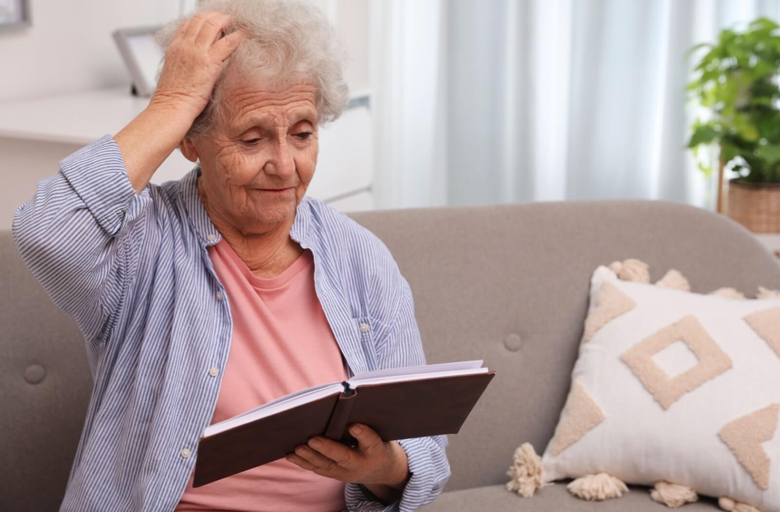 An older adult scratches their head while sitting on a couch and looking at a photo album in confusion
