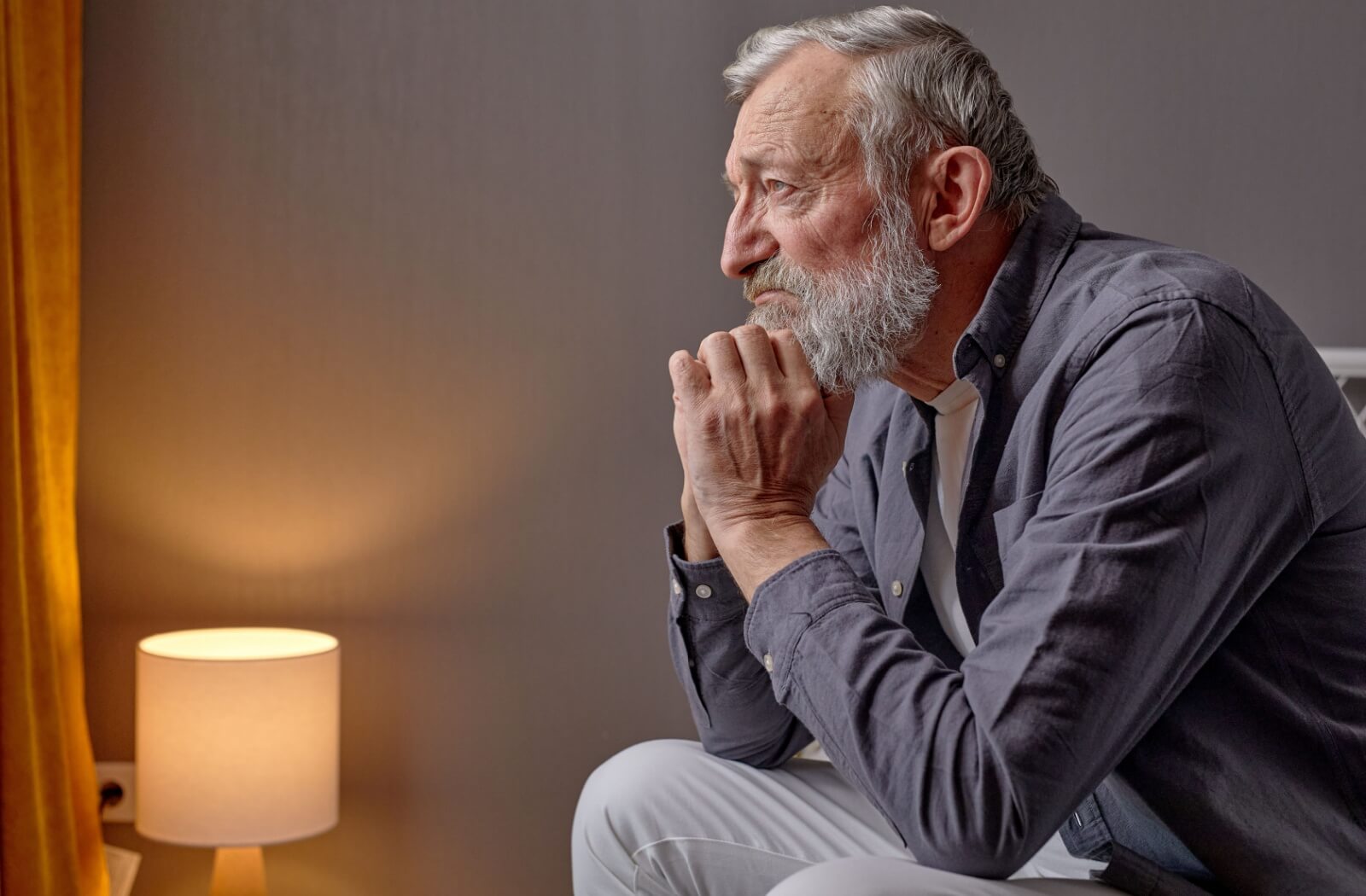 A sad looking senior man looking at a distance while he sits on his bed.