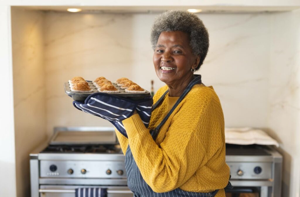 A happy senior woman holding up a tray of freshly baked muffins.
