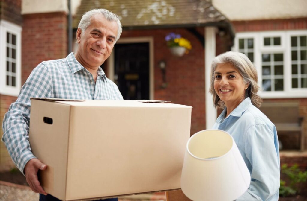 An older man holding a moving box and an older woman holding a lamp smiling at the camera