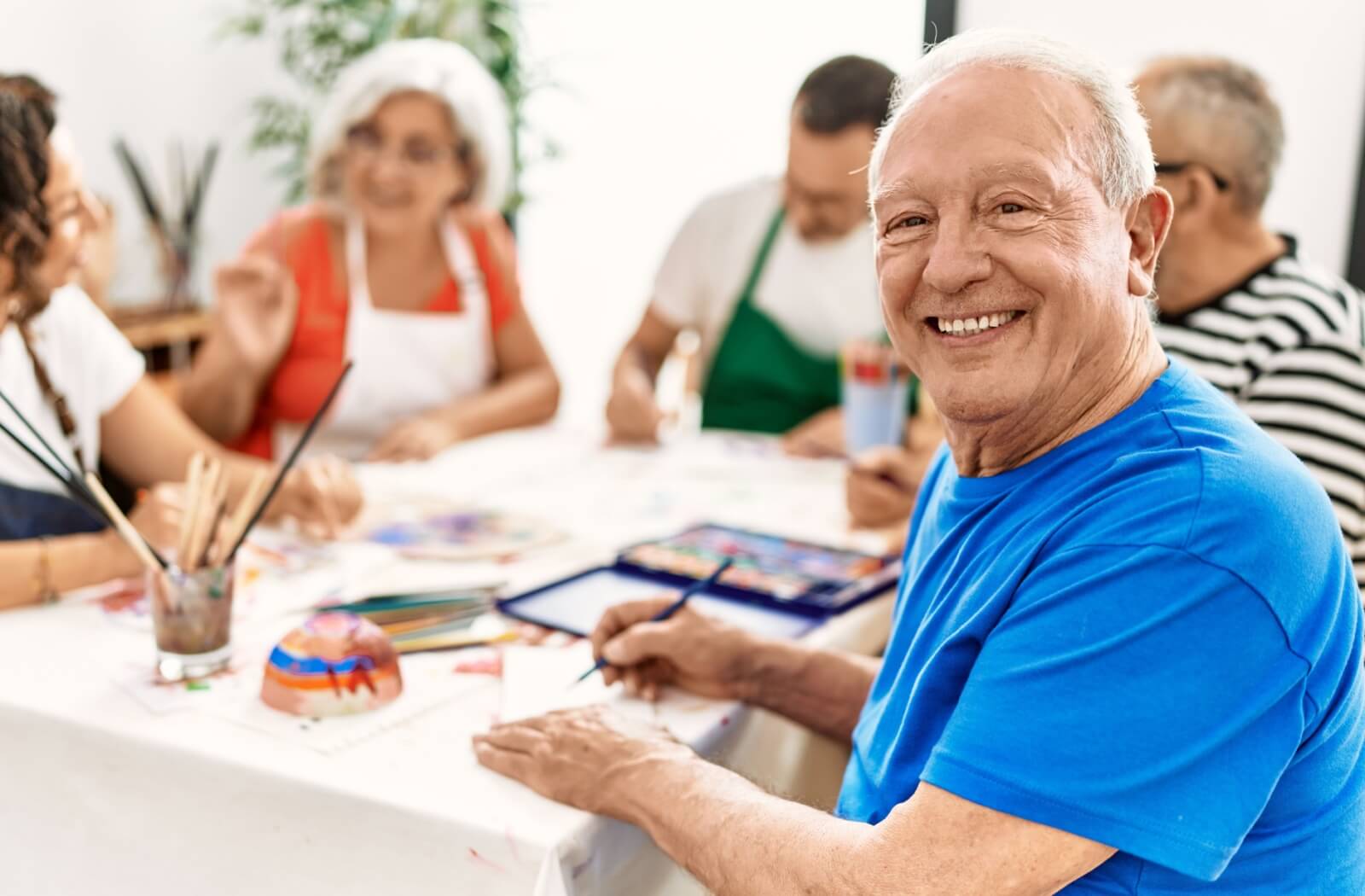 A group of older adults sits around a table, smiling and enjoying a group art class.