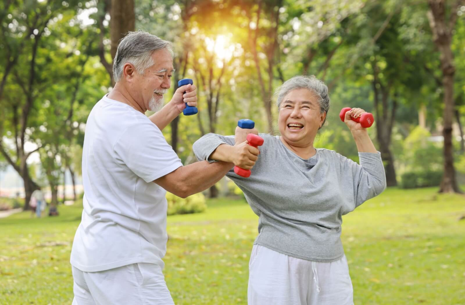 A senior couple playfully wrap their arms together while holding small dumbbells in the park to strengthen their bones