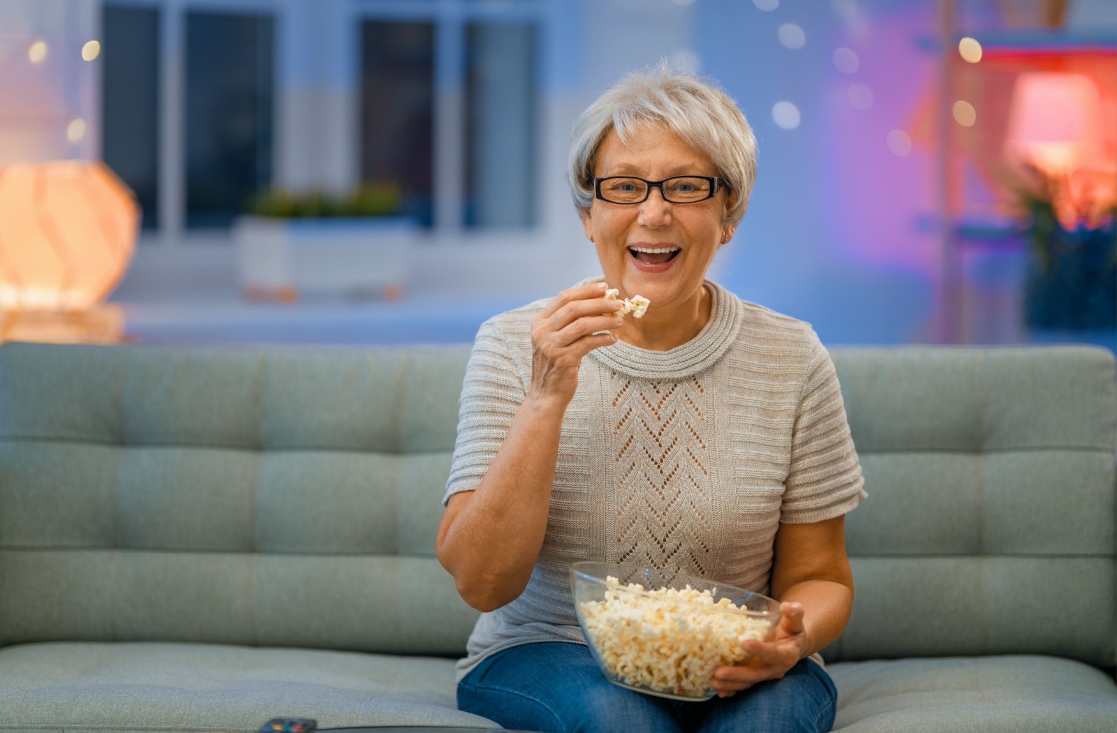 A senior resident enjoys a bowl of popcorn while watching a movie in their apartment at a senior living community.